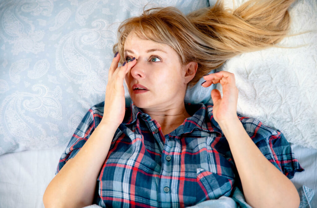 A woman lying in bed with pillows behind her rubbing her dry eye.