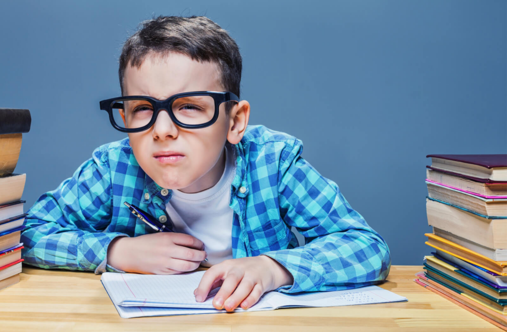A young boy with large black glasses is squinting and looking ahead while surrounded by books and writing in a notebook.