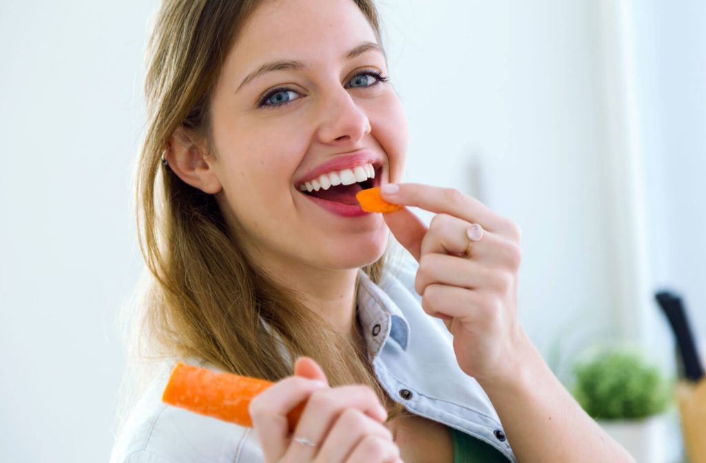 A young woman smiling, eating a carrot and looking directly in the camera