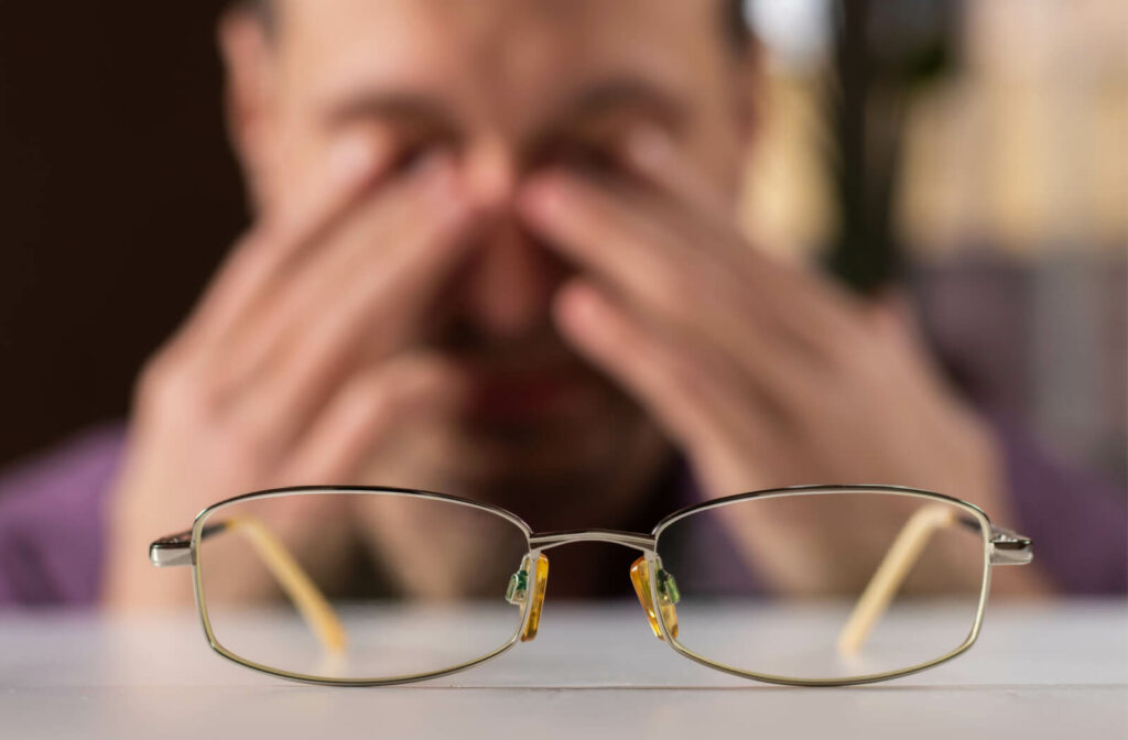 A young man is experiencing eye strain and discomfort, holding his glasses and rubbing his tired, dry, and irritated eyes due to prolonged computer work.